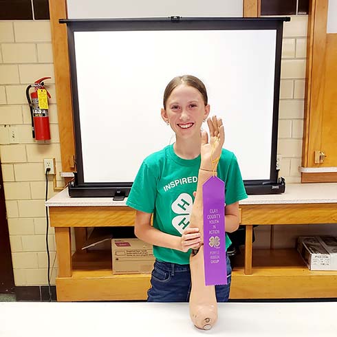 Chloe holding a fake arm with a 4-H ribbon hanging from the pinky finger